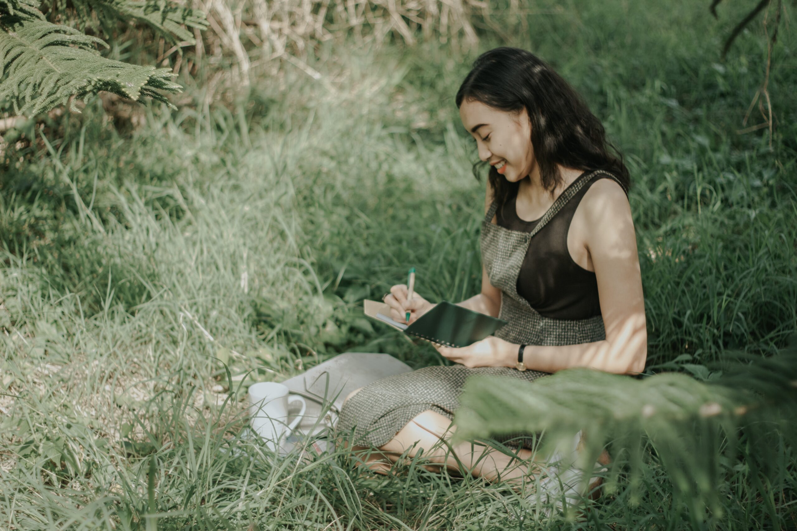 An Asian woman smiling and writing in a journal in a field.