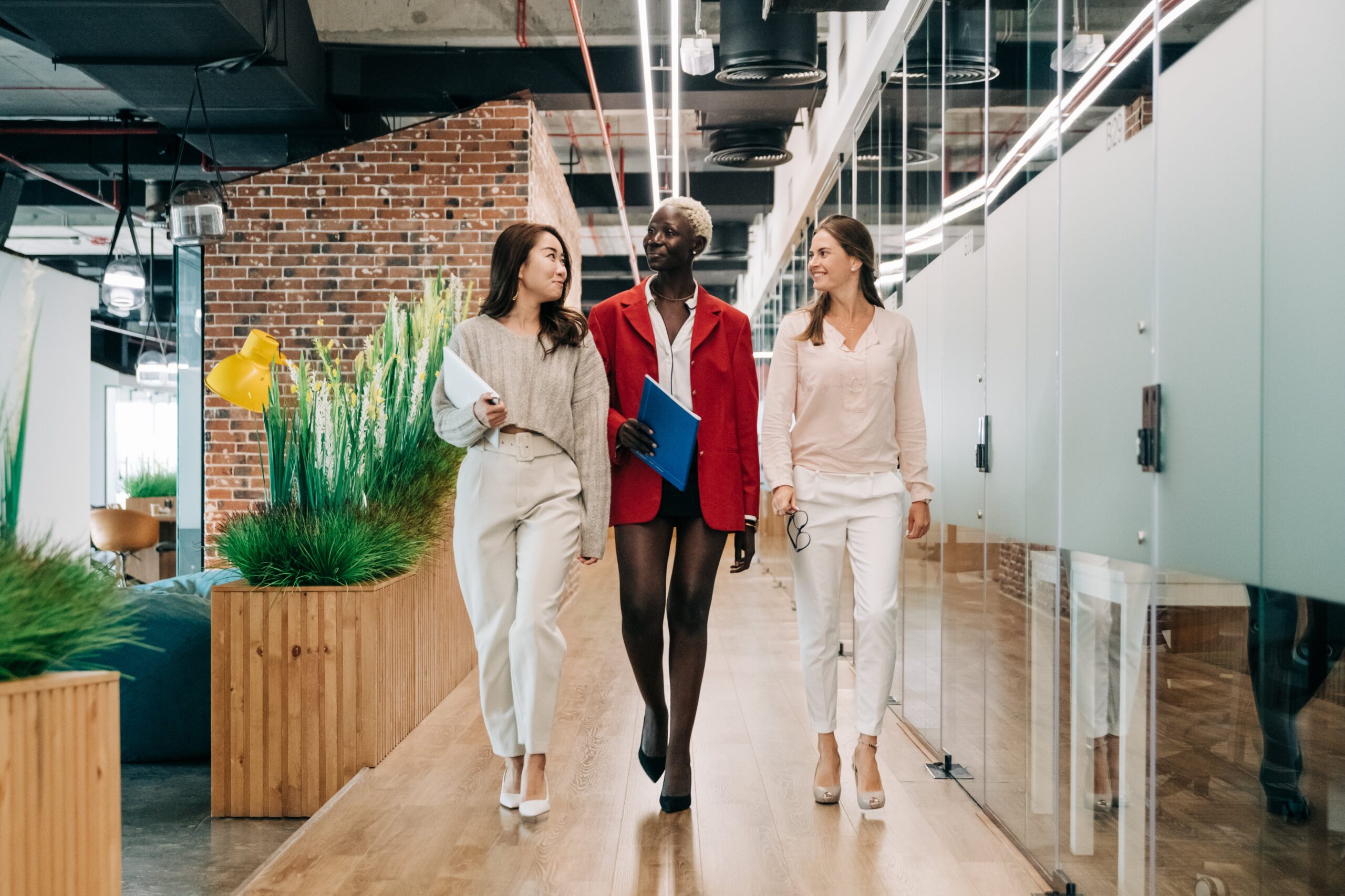 How to be an effective, empowering new manager. Here is a group of 3 women in the workplace, talking in the hallway.